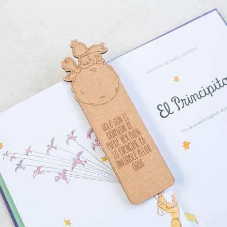 Detalle de marcapáginas de El Principito hecho de madera con frase y los personajes de el zorro, el Principito y la rosa grabados, situado en una página del propio libro