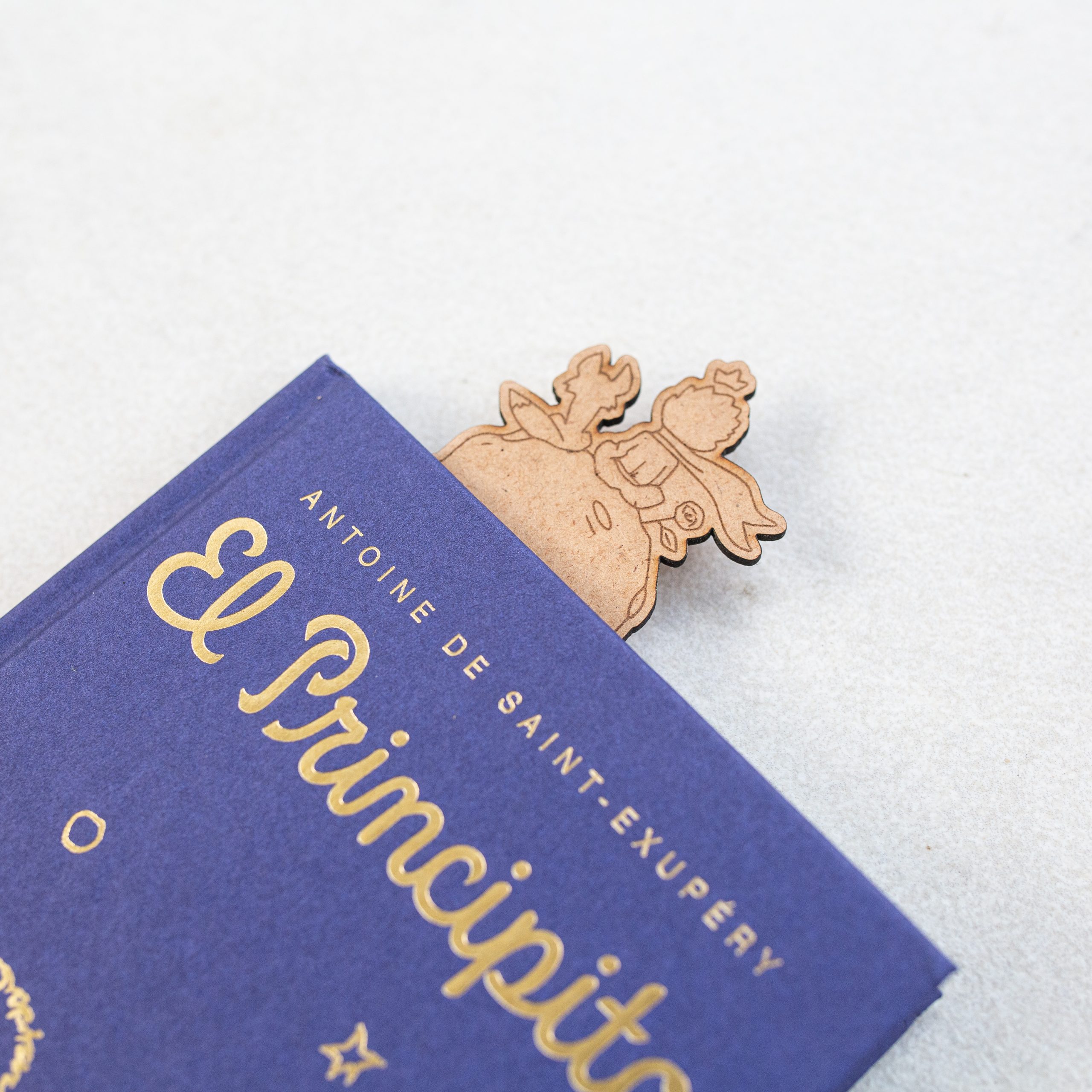 Marcapáginas de madera reciclada de El Principito para amantes del libro Detalle del libro El Principito con marcapáginas de este mismo libro en su interior con personajes de el zorro, el Principito y la rosa grabados en él.