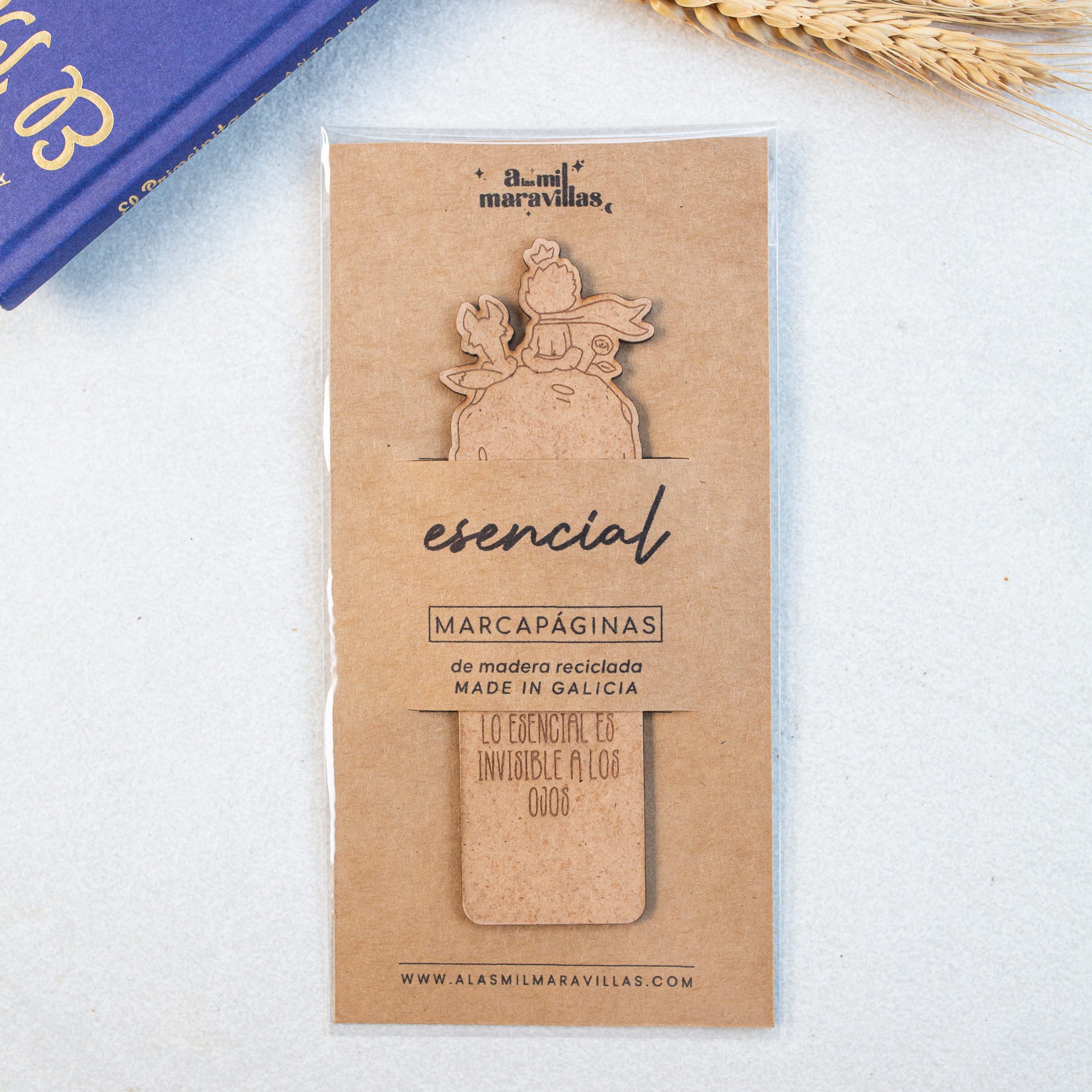 Marcapáginas para amantes de El Principito con personajes grabados Marcapáginas de El Principito hecho de madera en envoltorio de A las Mil Maravillas situado sobre superficie blanca con libro y hojas de trigo