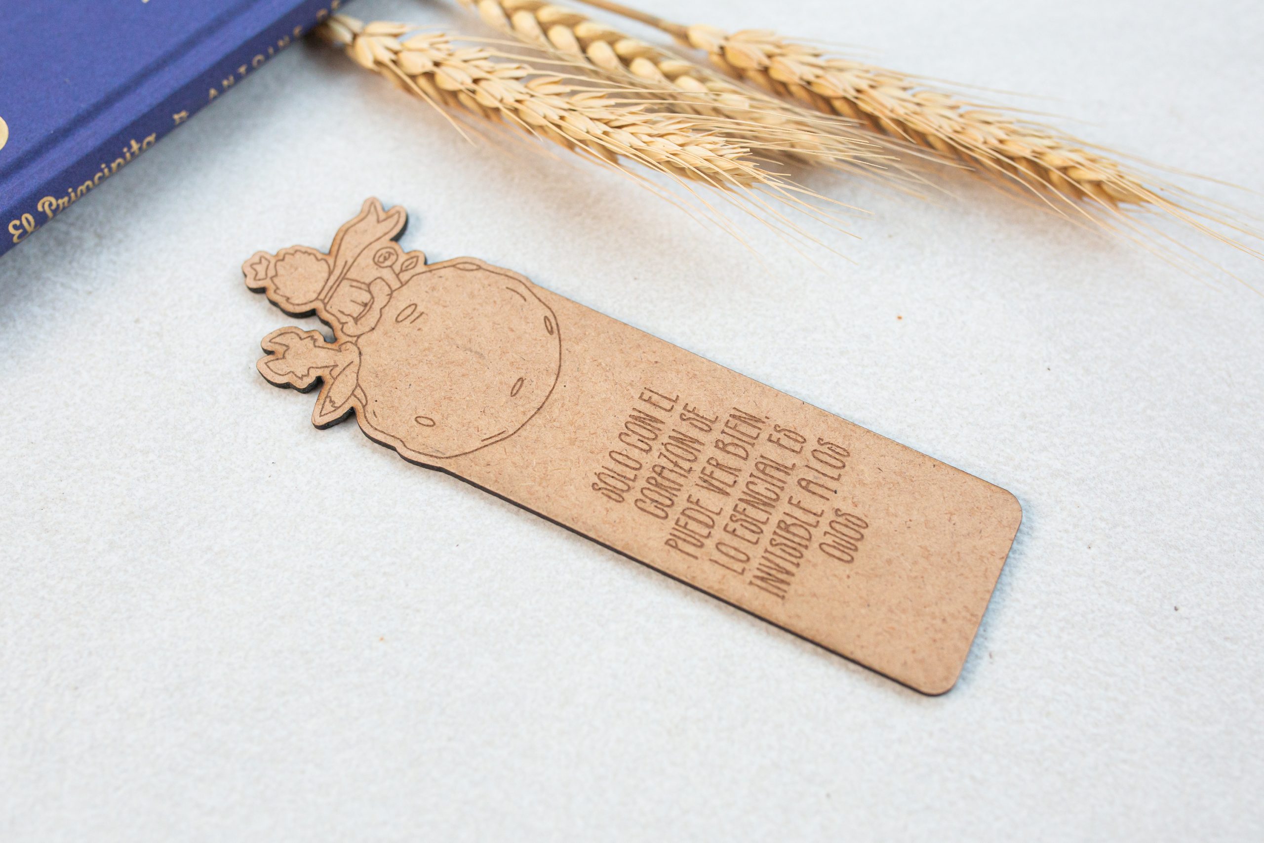 Marcapáginas de madera reciclada de El Principito Marcapáginas de madera de El Principito con dibujo de personajes y frase grabados. El marcapáginas está apoyado de forma casi horizontal sobre superficie blanca con libro de El Principito y tres hojas de trigo en una esquina
